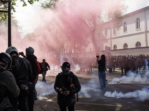 Manifestation urbaine sous un nuage de fumigène rose, journalistes casqués au premier plan